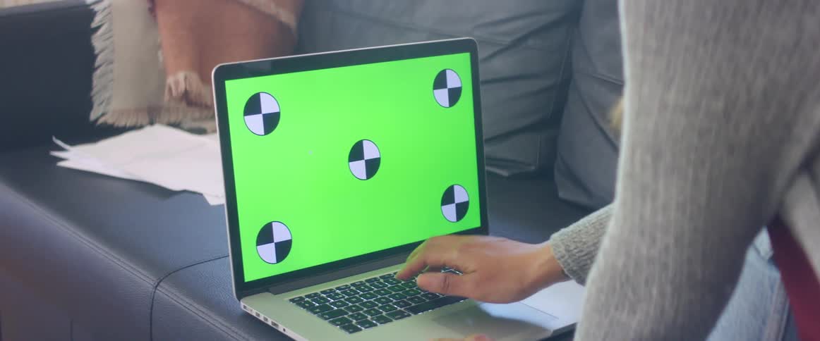Woman with jeans and a grey sweater sits on dark blue sofa uses laptop with greenscreen