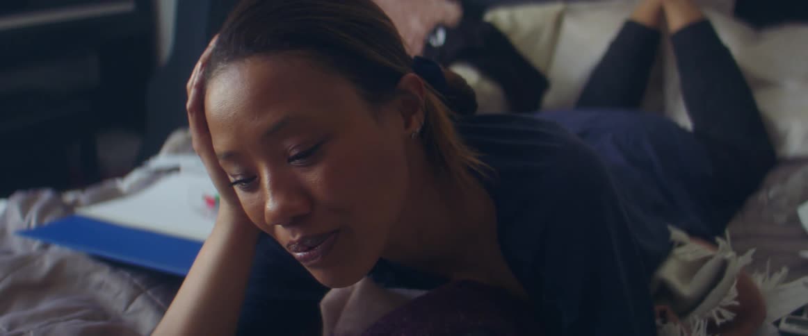 Woman with earrings and a ponytail lies on bed and looks at laptop smilingly in the bedroom