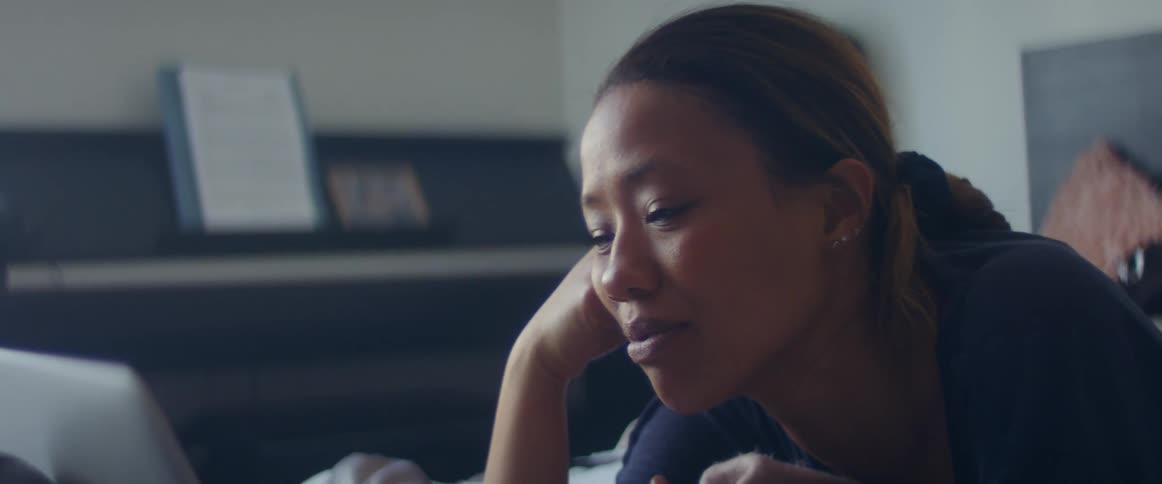 Woman with earrings and a ponytail lies on bed and looks at laptop in the bedroom