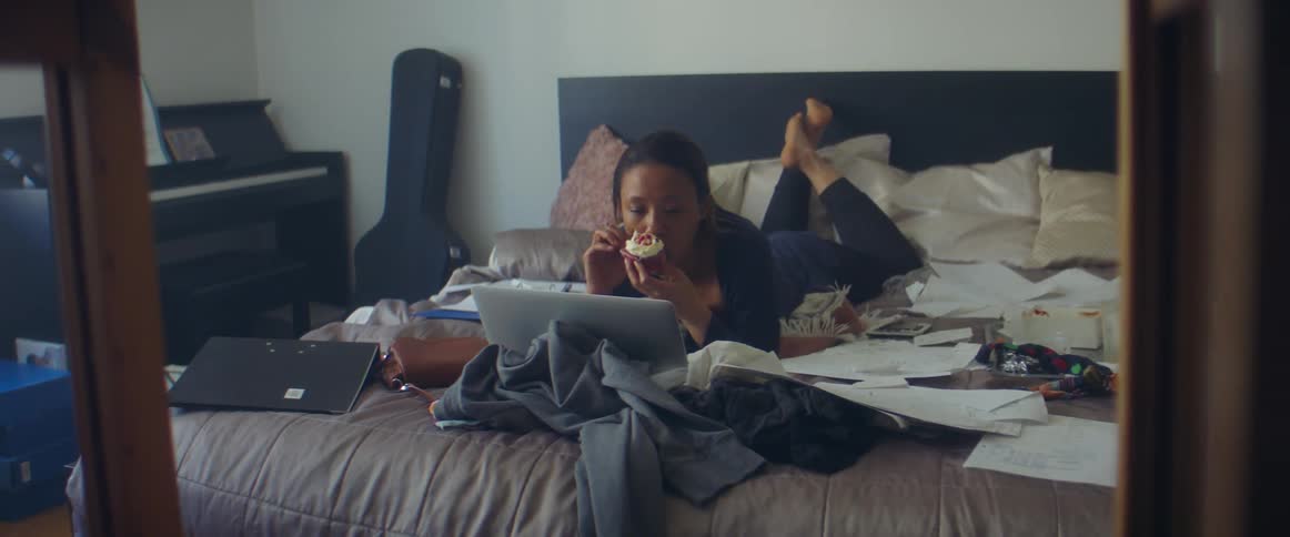 A woman takes a bite from a cupcake while looking at a laptop as she lies on her stomach on a bed covered in paper and clothing