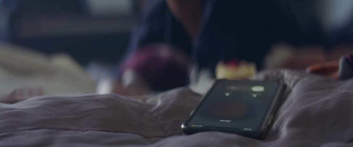 A woman in a bedroom laying in bed with a blanket with a smart phone ringing