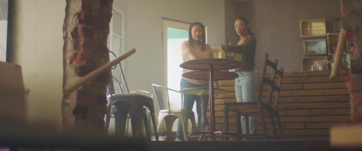 Two women in a room moving tables and chairs with a wall and doorway in the background