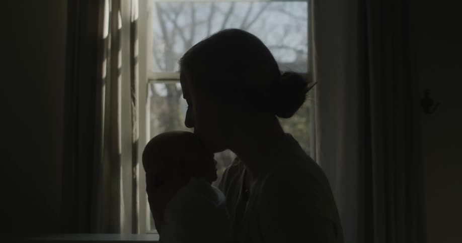 A woman picks up and holds her newborn while standing in front of a window