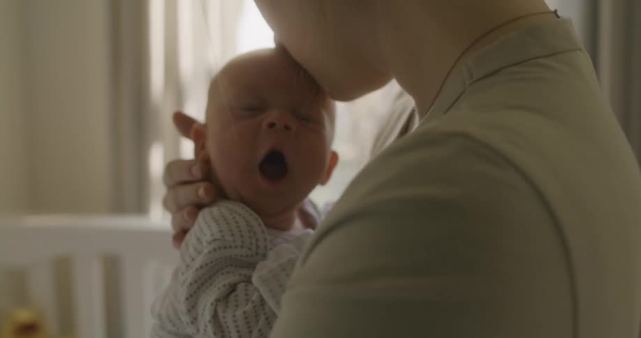 Mother holding a yawning baby in her arms inside a room at home