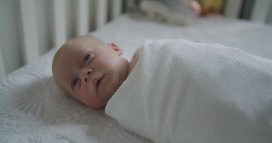 Newborn baby wrapped in a white blanket resting on a crib in a room at home