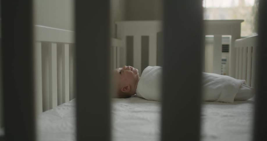 Baby swaddled in blanket lies on his back on mattress in crib near window