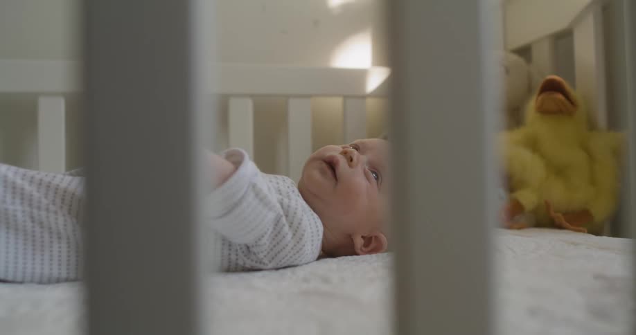 Baby lies down in crib near stuffed toys waving arms and smiling