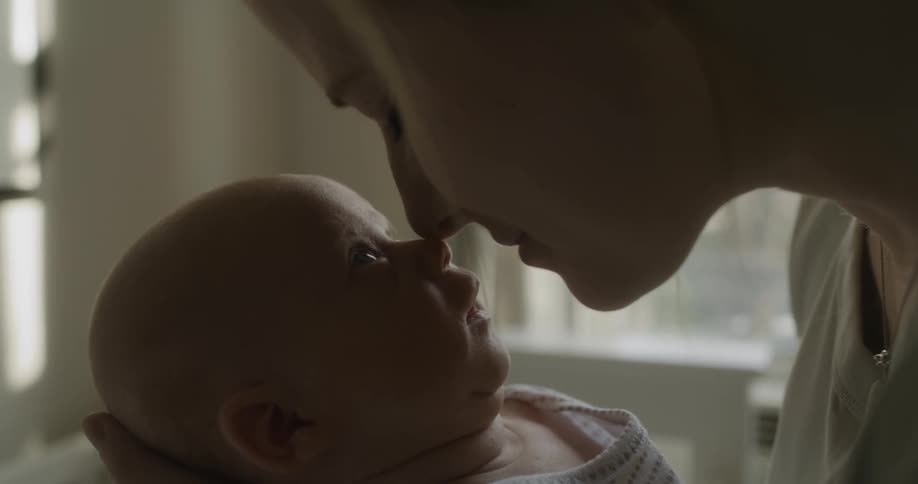 Mother holds baby rubbing noses together while smiling standing near crib set near window