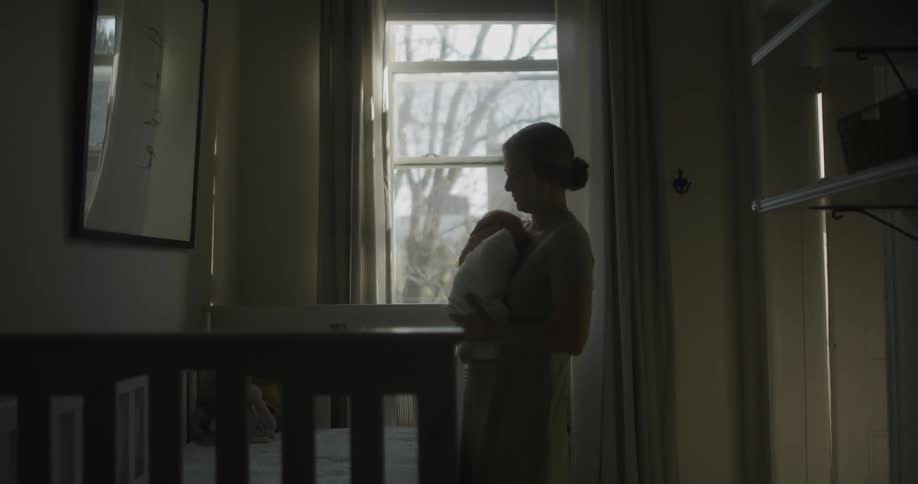 A woman picking up a baby from a crib with a wall and window in the background