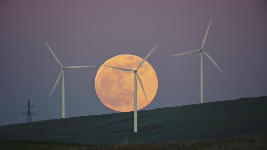 A super moon rises in a purple sky with three wind mills and a high voltage transmission line in the foreground in this unreal green energy shot