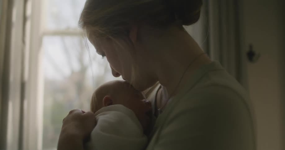 Blonde woman with a brown shirt holds and kisses her baby next to window in baby room