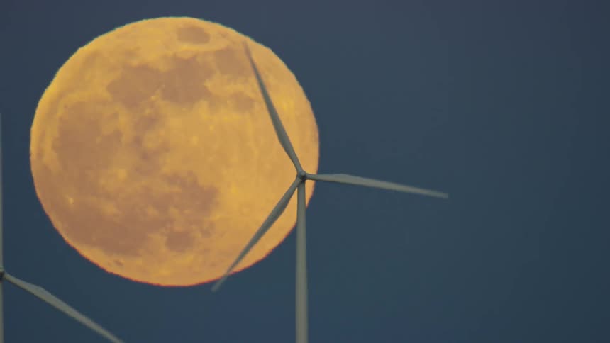 Tight shot of a golden super moon rising with two wind mills generating renewable green energy in the foreground with a post sunset blue sky in the background