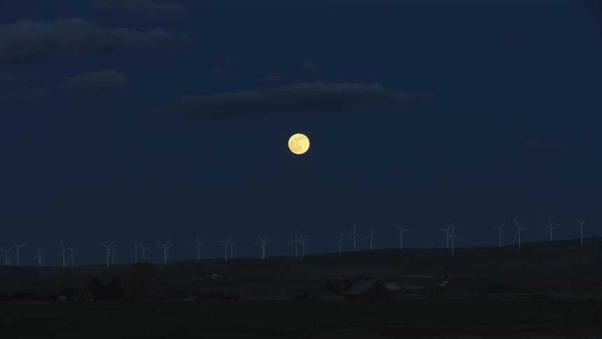 Blue hour post sunset tracking shot of a ridge of wind turbines generating clean renewable electricity with a full super moon center framed in the sky above communicating balance and evenness visually