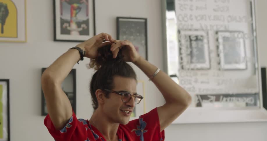 Man puts his hair up into a pony tail on top of his head standing next to wall of pictures while talking and smiling
