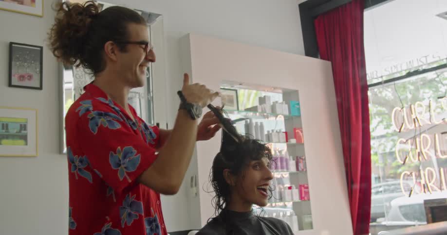 Male hair stylist cuts womans hair as they talk in room with view of business sign and cars