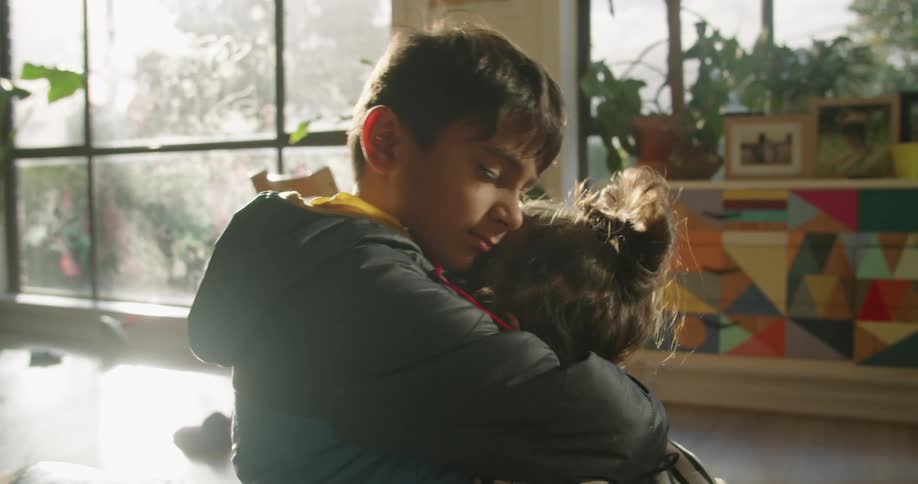 A toddler smiles as she walks up to her older brother and gives him a hug