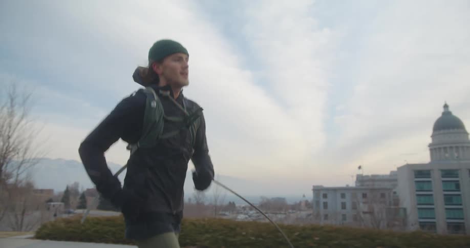A man with a hat jogging with buildings trees the sky and clouds in the background