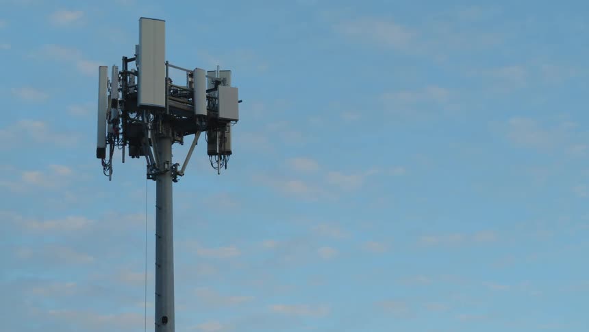 Orbiting close below antennas mounted to a cell tower with blue sky and whispy clouds in the background