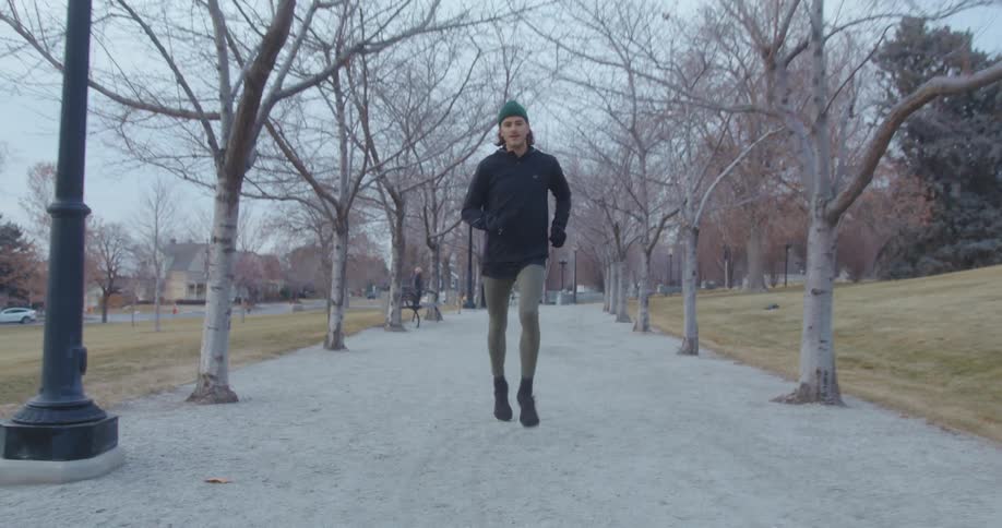 Man wearing cold weather gear jogs on track in park as car drives on street near apartments nearby