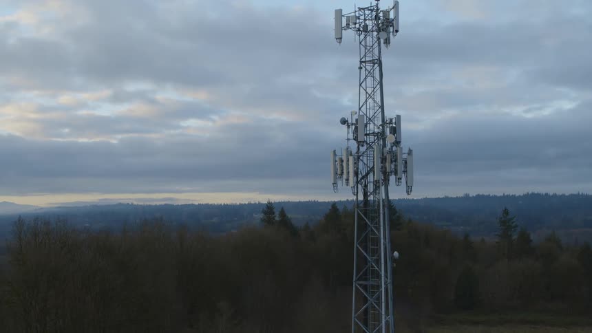 Pullback reveal of a cell tower that rises high above a forest and trees with mountains in the distance