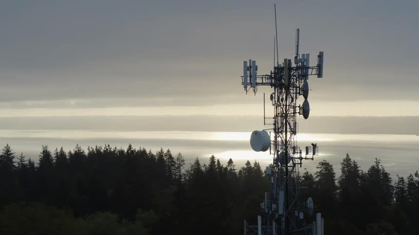 Orbit around a backlit 5G data tower high above the trees with a calm coastal bay in the distance