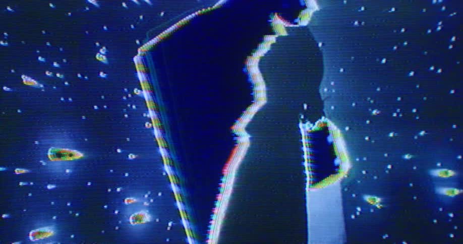 A digital display features a man standing at profile in front of a spinning cube and projectiles