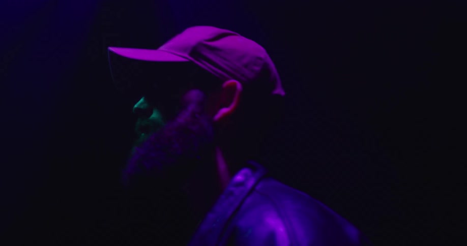 A man stands in a baseball cap and leather jacket under color changing neon lights