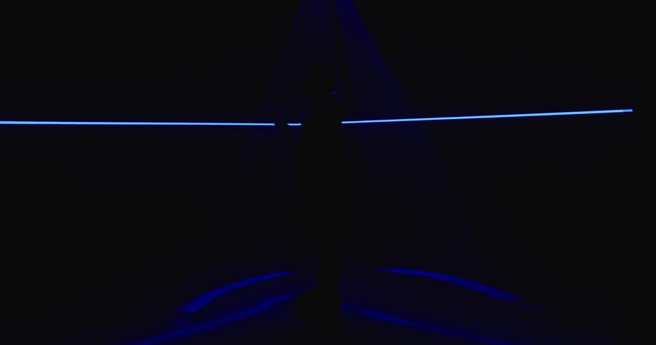 Man stands in middle of dark room lit by blue and red lights with neon line moving across wall as he twirls drum stick