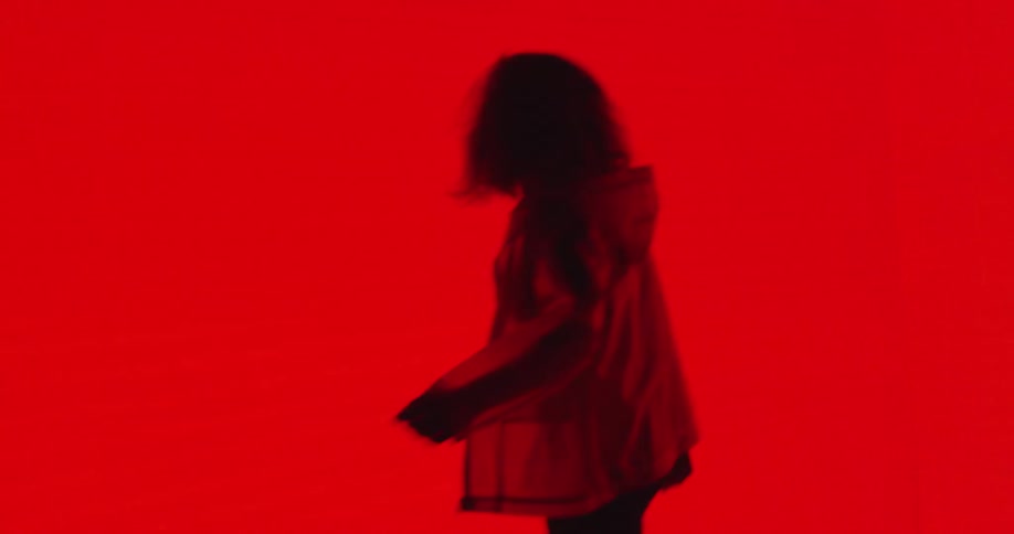 A woman dances around a room with glowing red walls in a mesh jacket and combat boots