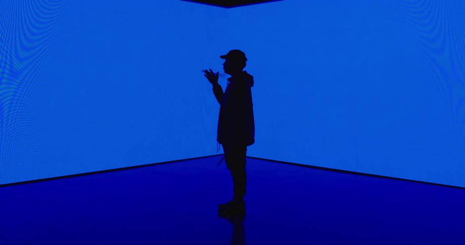 A silhouetted man spinning a drumstick in front a blue glowing wall