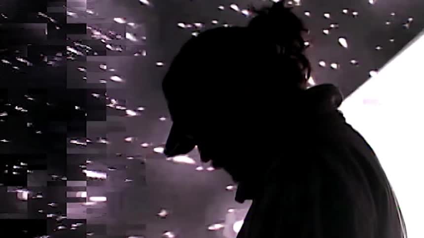 A man in a baseball cap and coat stands in profile in front of a giant screen with a spinning cube and exploding comets