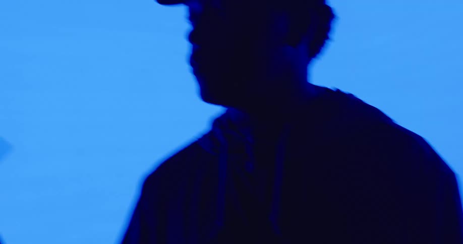 The silhouette of a man in a cap twirling a drumstick as blue lights and shadows surround him