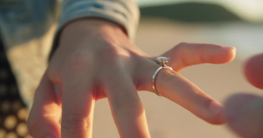 A man slides an engagement ring onto his girlfriends finger as they stand on the beach