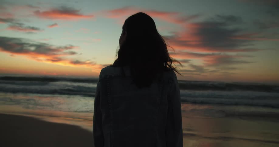 Woman standing and watching the sea on the beach is silhouetted against the red sky