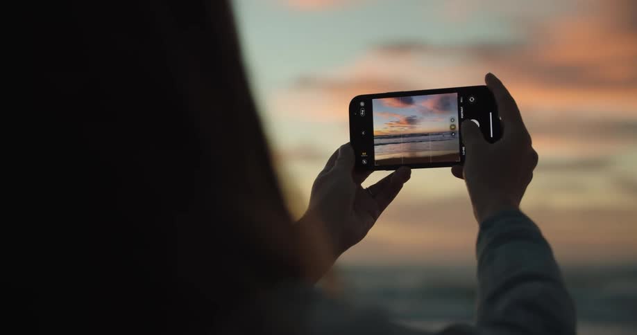 Woman takes pictures of the sunset over the ocean with a smartphone