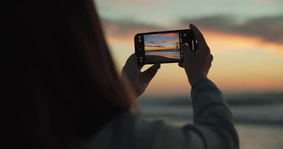 Woman with brown hair takes a photograph of sea with red sky on the beach at dusk