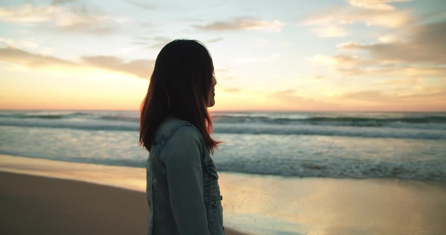 Sun sits on pink horizon over ocean as woman stands on beach looking out to sea