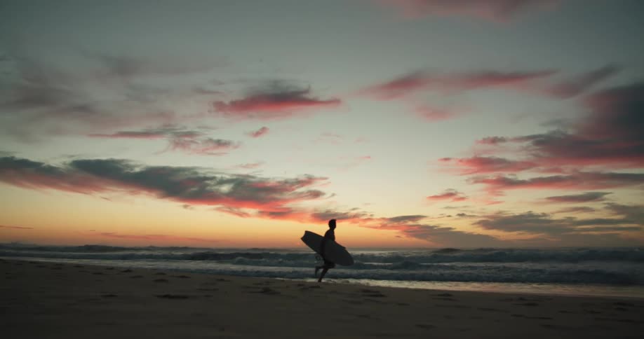 A silhouette of a person running with a surfboard on the beach with the sky and clouds in the background