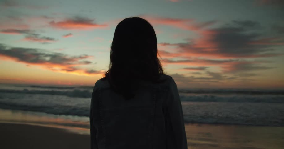 Woman standing and watching the sea on the beach is silhouetted against the red sky