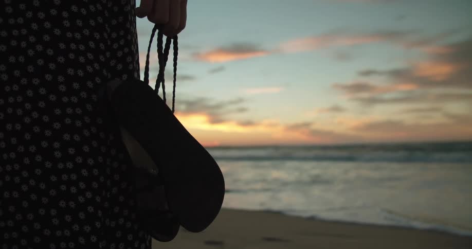 A female stands on the beach holding her shoes while looking out at the water