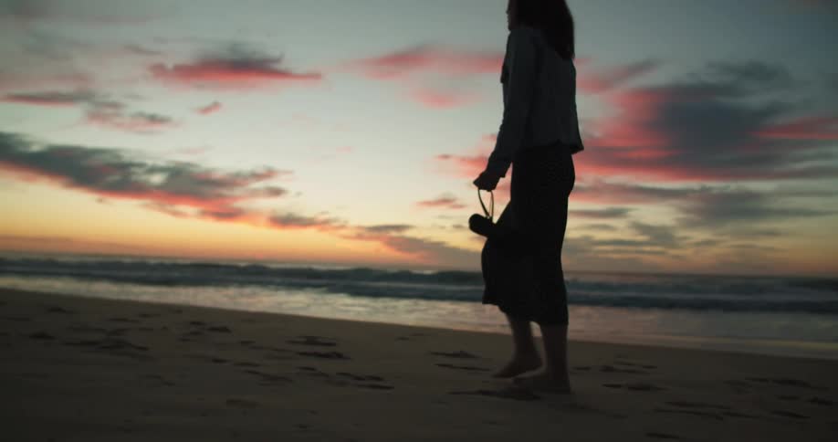 The silhouette of a woman carrying her shoes as she walks on the beach sand at sunset