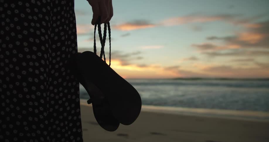A woman is walking on a beach towards the sea at sunset while holding her shoes