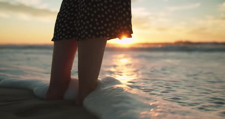 A woman standing in a beach with sand with the waves of water washing over her feet with the sky and clouds in the background