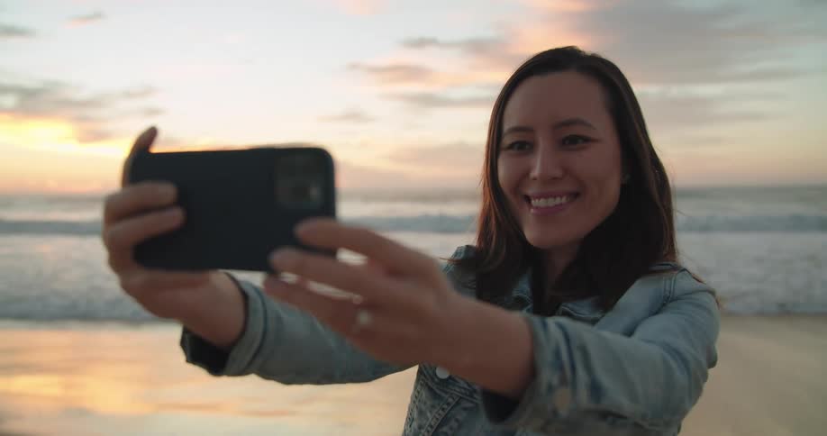 Woman with straight hair and a denim jacket takes a selfie with a smartphone on the beach next to the sea at dusk