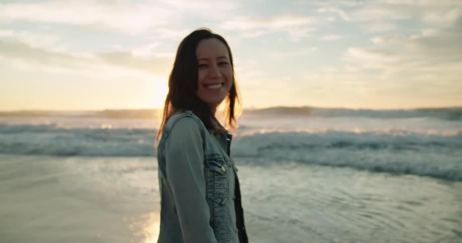 A woman in a dress and denim jacket smiles as she spins around on the beach