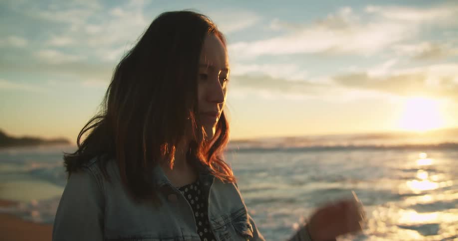 A woman stands near the ocean water with the sun low near the horizon and moves the hair from her face