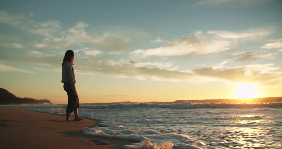 A woman stands on the beach alone looking towards the ocean water