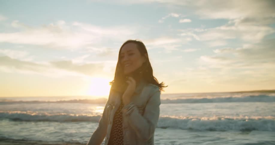 Woman with brown hair and a denim jacket turns around on the beach next to the ocean at sunset