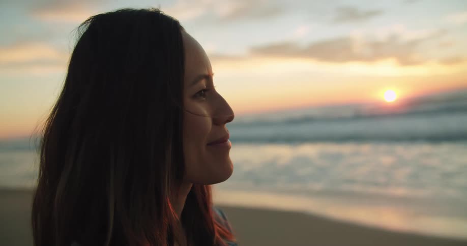 Brunette woman with brown hair smiles and looks around on the beach at sunset