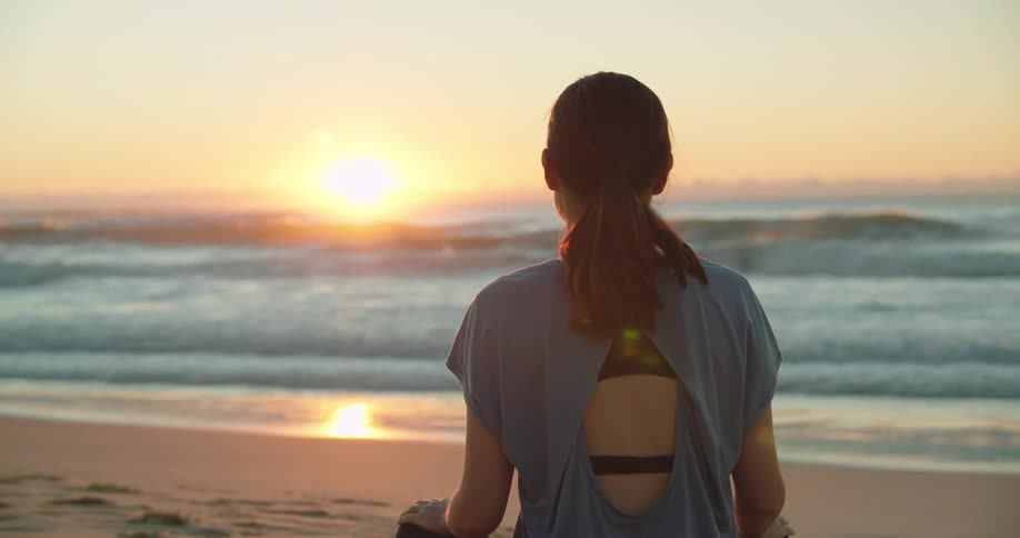 A woman sits cross legged on the sand looking out at the ocean waves at sunset
